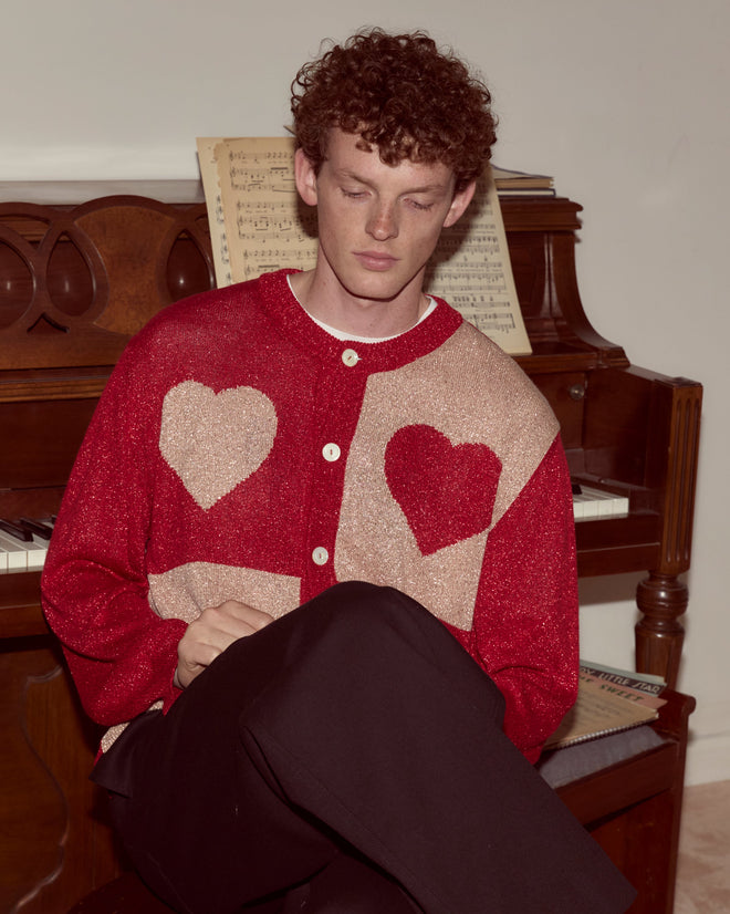 A man in a Lamé Hearts Cardigan sits by a piano with sheet music, capturing an Alice in Wonderland-inspired whimsical charm.