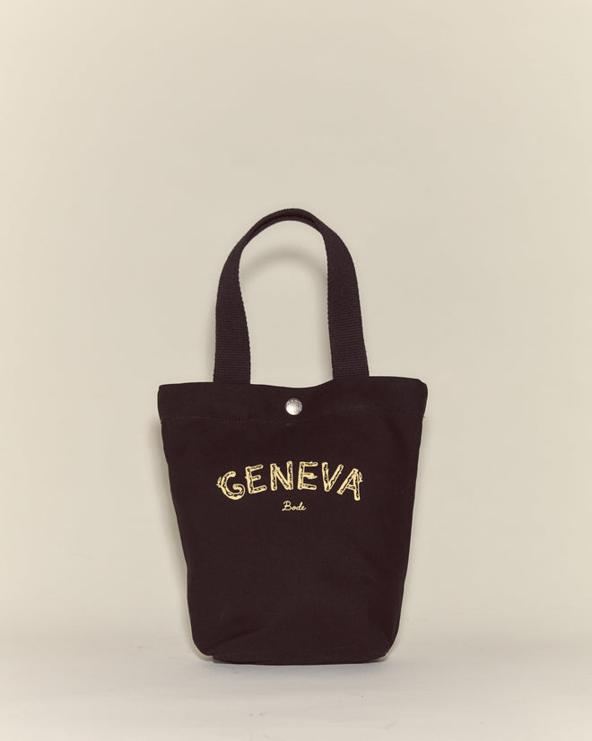 The Small Geneva Tote is a black canvas bag featuring "GENEVA Belle" embroidered in yellow thread, displayed on a beige background—a chic accessory inspired by Camp Geneva.