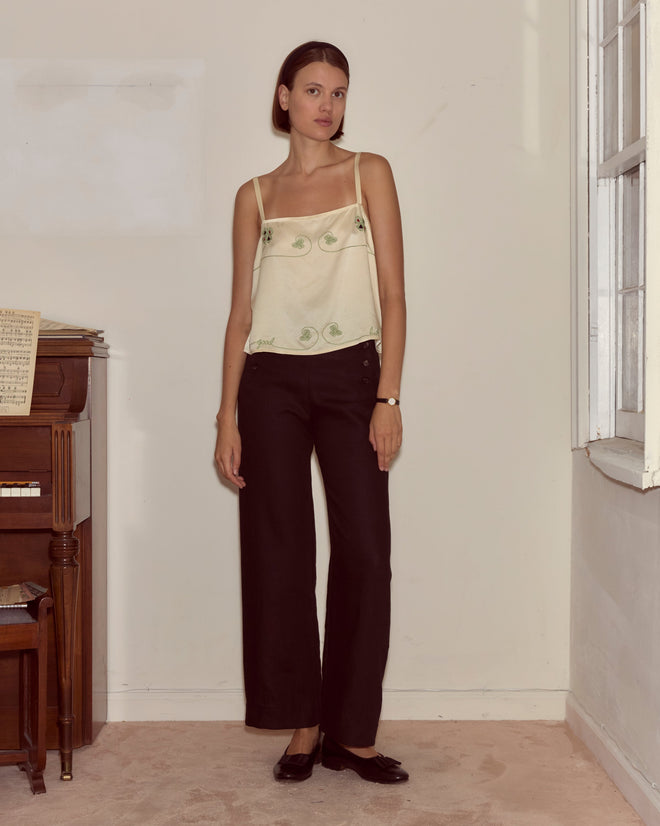 A woman wearing the Lucky Draw Tank—hand-embroidered silk with a subtle playing card motif—and black pants stands indoors by a piano and window, looking at the camera.