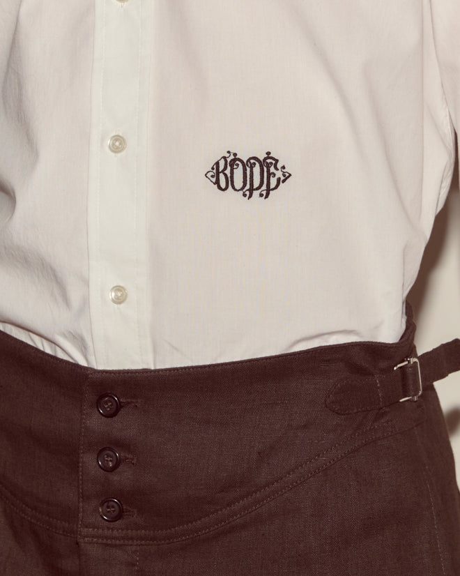 A Monogrammed Poplin Shirt in white, made from 100% cotton with "BODE" embroidered on the chest, styled with high-waisted brown pants.