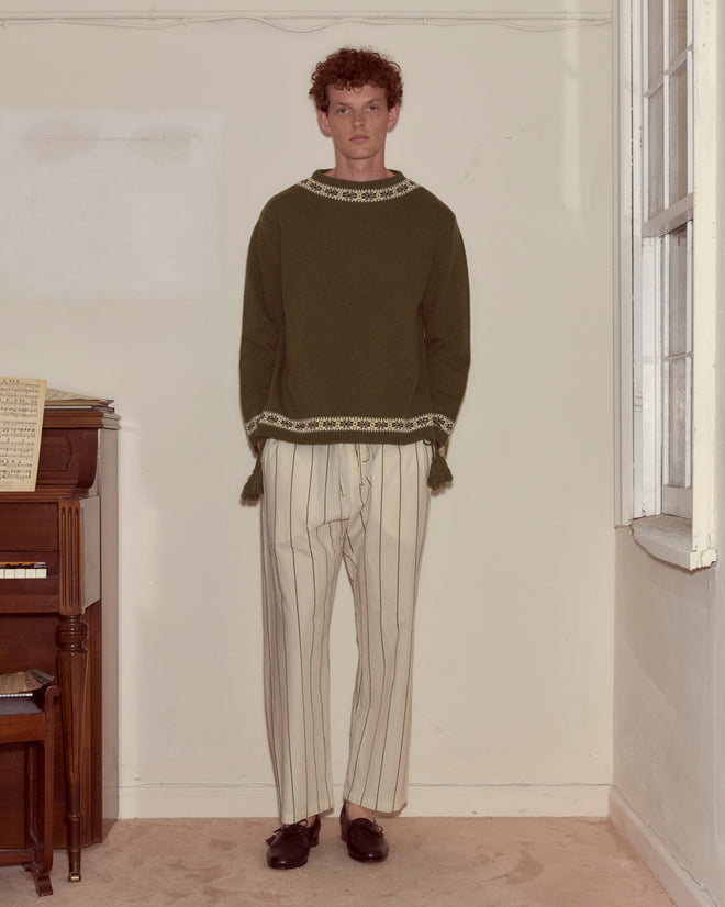 Indoors by a piano and window, a person wears the Moritz Sweater, crafted in green virgin wool, paired with white striped pants.