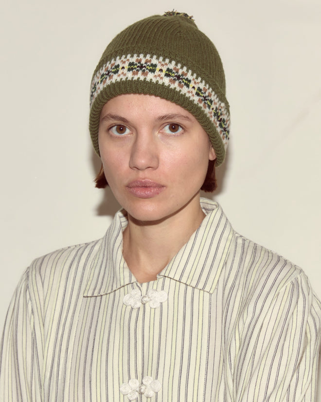 Person wearing a green Moritz Hat made of merino wool in a Fair Isle pattern and a striped buttoned shirt, looking at the camera with a neutral expression.