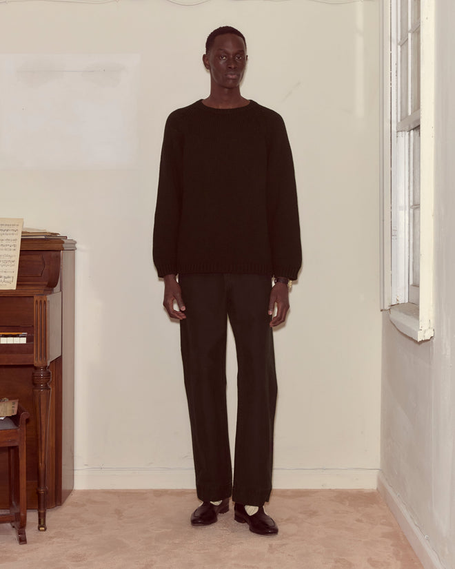 A person wearing the Cashmere Crewneck - Black stands indoors beside a piano and a window.