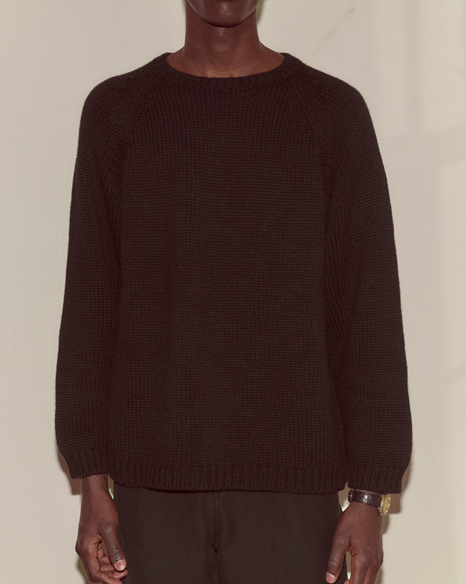 A person wearing the Cashmere Crewneck - Black with an oversized fit stands against a beige wall, arms relaxed at their sides.
