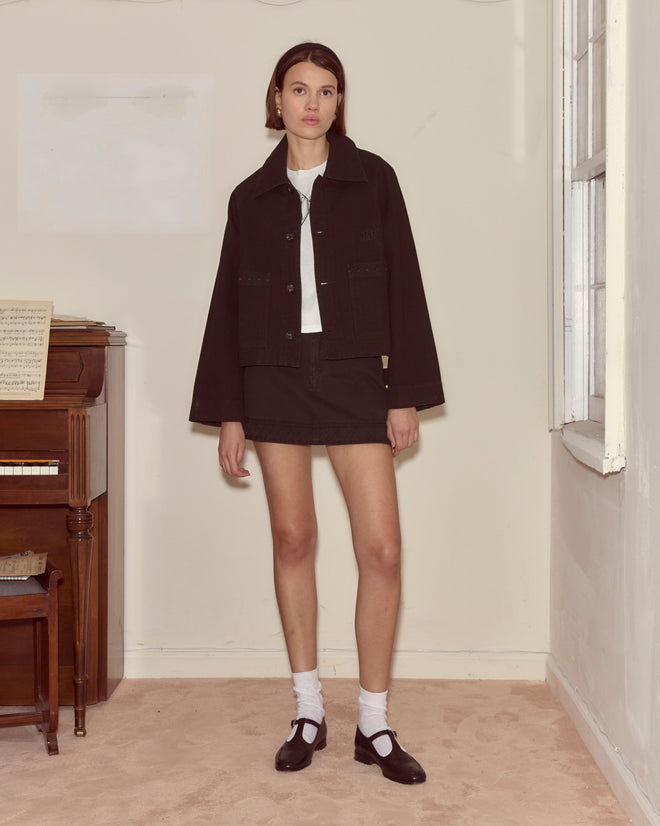 A woman in an embroidered black denim jacket stands indoors by a piano, paired with a skirt, white socks, and black shoes.