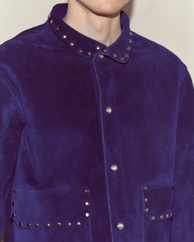 Person wearing the Studded Welding Jacket - Blue, featuring ultramarine suede with metal studs on the collar and pocket flaps, inspired by classic welder's jacket style.