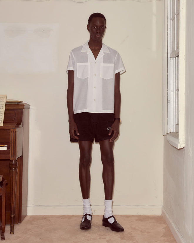 Wearing the Voile Short Sleeve Shirt - White and black shorts, a person stands indoors by a window next to a piano.