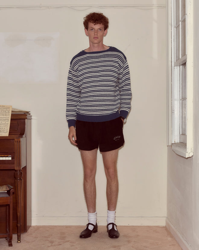 Indoors, a person wears the Peter Stripe Sweater from the latest men’s knitwear collection, styled with black shorts, white socks, and black shoes.