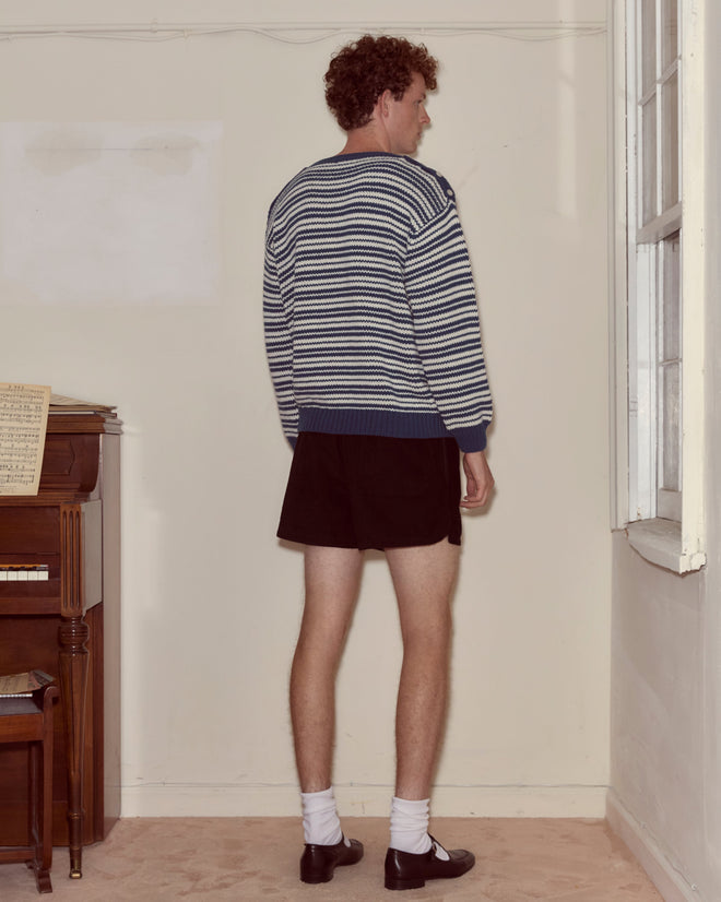 A person wearing the Peter Stripe Sweater, black shorts, and dress shoes stands facing a wall in a room with a piano.