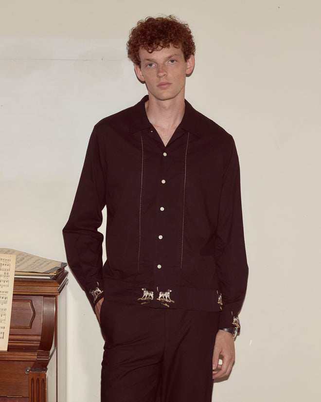 A man wearing the Pointer Long Sleeve Shirt in black, featuring subtle ladderwork details, stands by a piano with sheet music against a plain wall.