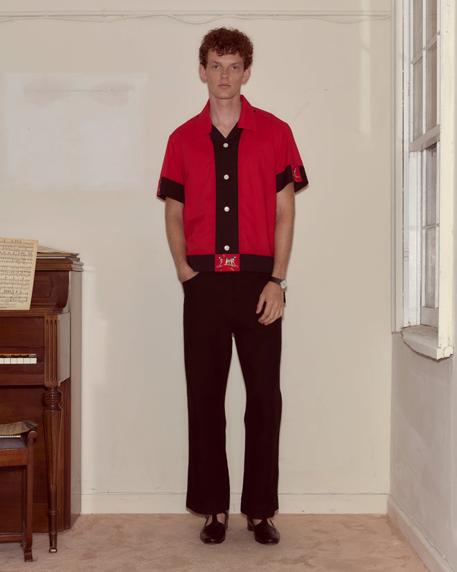A man in the Pointer Short Sleeve Shirt, featuring red and black cotton with subtle ladderwork detailing, paired with black pants, stands by a piano in a simple room with beige carpet.