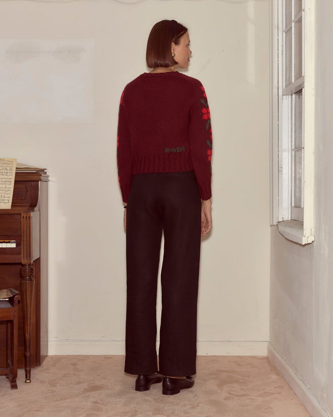 A woman with short brown hair stands facing away, wearing a maroon Skunk Sweater and black pants in a room with a piano.