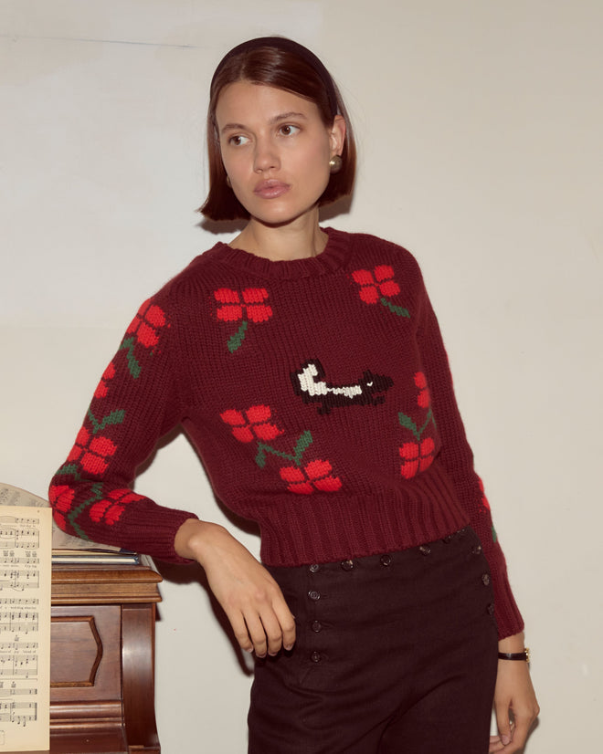 A woman in the Skunk Sweater, a chunky red floral knit, leans against a piano with sheet music, gazing off to the side.
