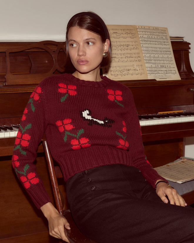 A woman wearing the Skunk Sweater—a chunky red knit with floral patterns—sits by a piano with sheet music in the background.