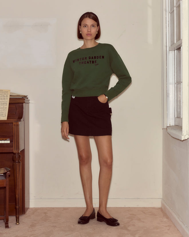 Woman in a dark green Theatre Crewneck and black skirt stands by a sunlit piano, channeling Broadway charm with a nod to Peter Pan.