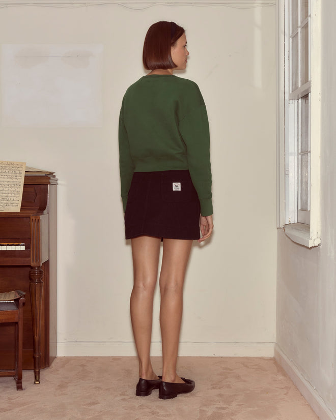 A woman stands indoors facing a wall, wearing the Theatre Crewneck - Dark Green with a black skirt and black shoes—a look inspired by the vibrant energy of Broadway.