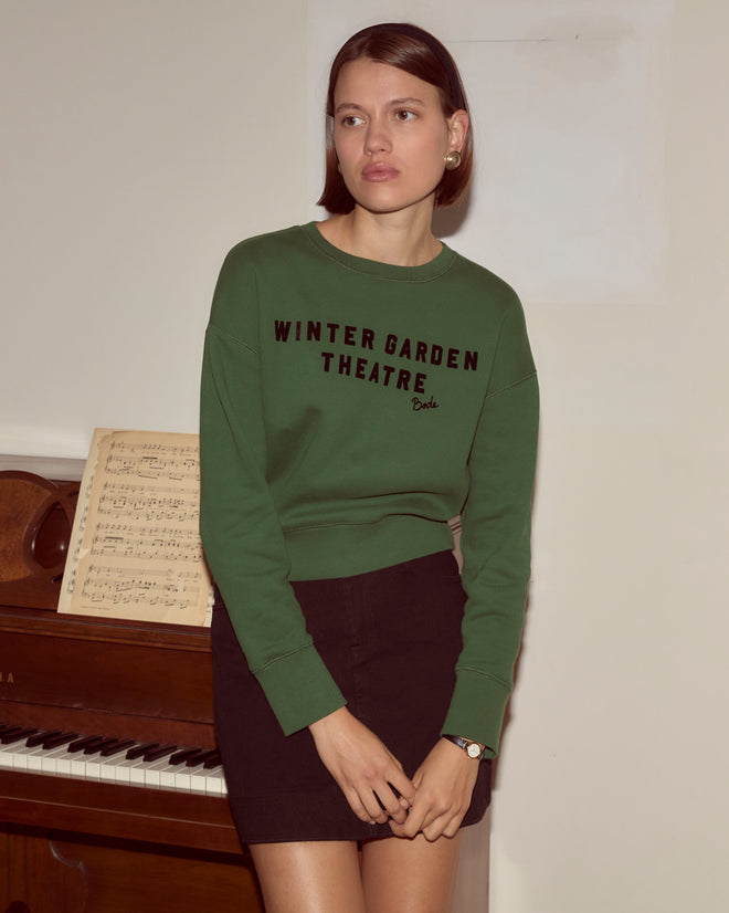 A woman in a dark green Theatre Crewneck stands by a piano with sheet music, evoking Broadway magic—perhaps inspired by Peter Pan.