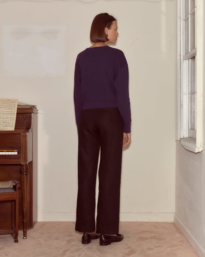 A woman stands indoors by a window and piano with sheet music, wearing the Theatre Crewneck in dark purple.