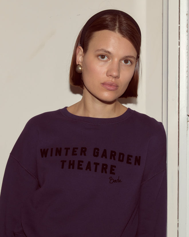 Woman with straight brown hair and headband wearing a dark purple "Theatre Crewneck," facing the camera.