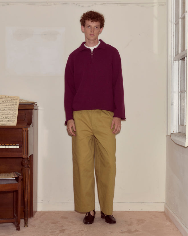 Someone wearing the Wool Dock Pullover, a chunky maroon oversized sweater, and tan pants stands indoors beside a piano and a large window.